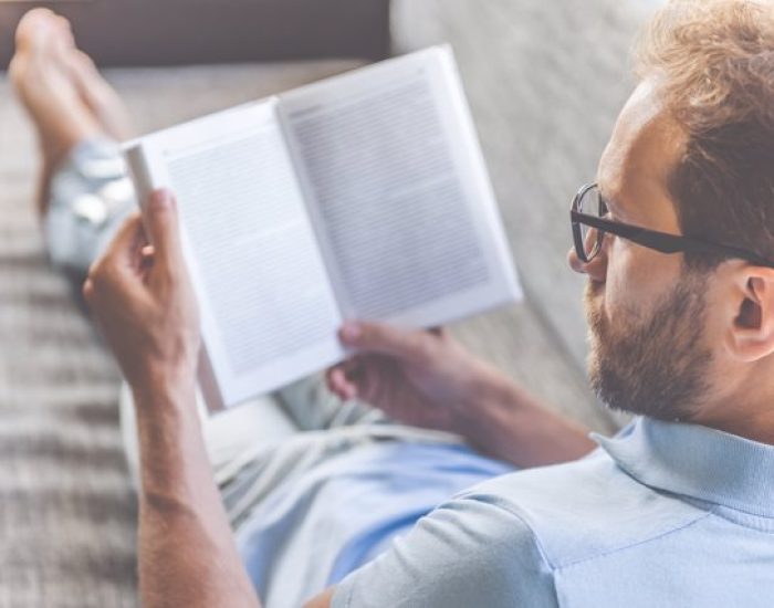 Back view of handsome young businessman in casual clothes and eyeglasses reading a book while lying on couch at home