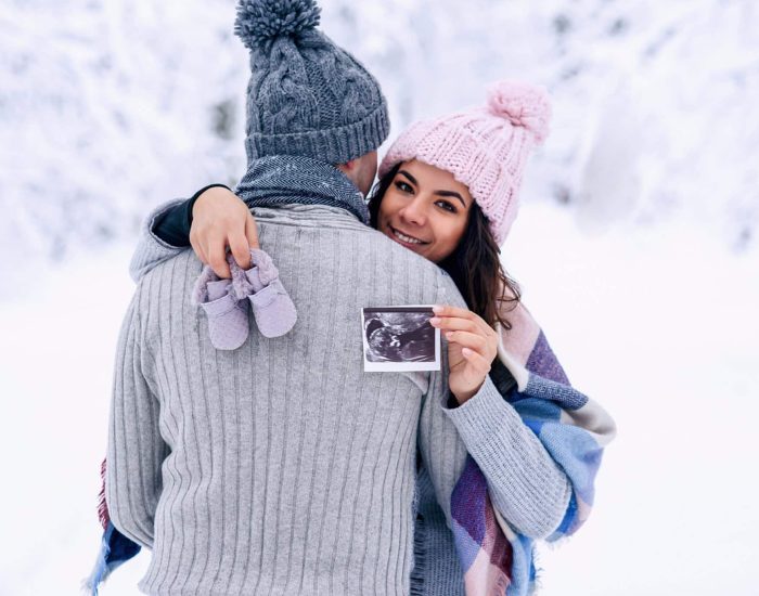 Beautiful pregnant woman in warm pink hat holds ultrasonic pregnant picture and baby shoes in her hands behind his husband back. Family love and care concept.