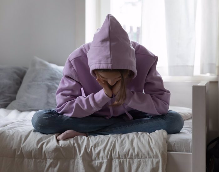 Unhappy depressed teenage girl wearing sweatshirt sitting on bed alone, feeling lonely and misunderstood, stressed sad teenager thinking about troubles, hiding face, child and psychological problem