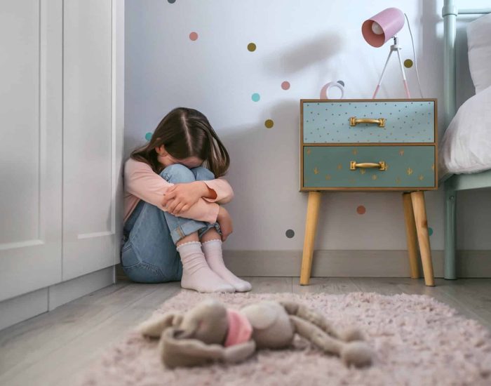 Unrecognizable sad little girl sitting on the floor of her bedroom with stuffed toy lying. Selective focus on girl in background