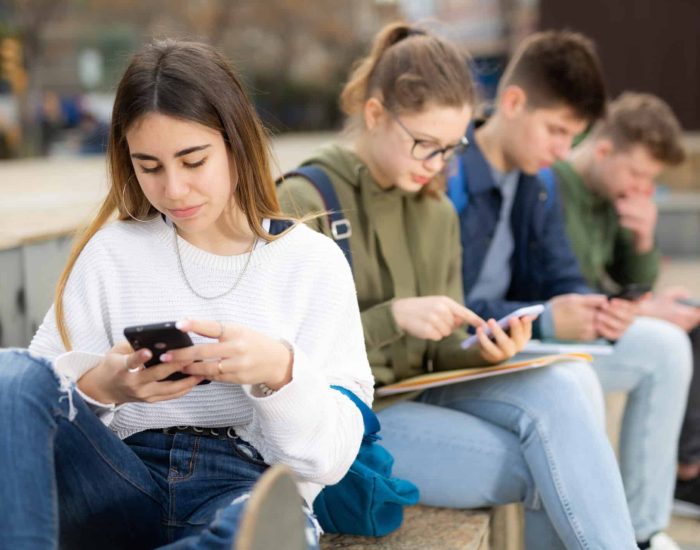 Portrait of girl teenager addicted in phone sitting with friends on bench outdoors