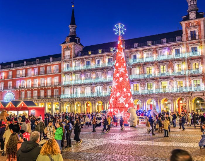 Madrid, Spain - December 18, 2023. Festive crowd gathers in Plaza Mayor at nightfall, illuminated by a shining Christmas tree with the main facade of the Casa de la Panadería in the background. Madrid, Spain.