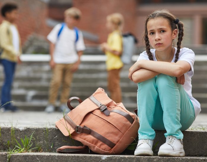 Full length portrait of sad schoolgirl looking at camera while sitting on stairs outdoors with group of children in background, copy space